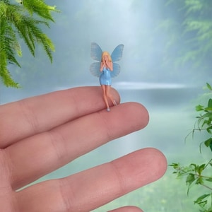 May include: A miniature fairy figurine with blonde hair, a blue dress, and translucent blue wings. The fairy is standing on a finger, with a blurred background of a lake and greenery. A whimsical decorative item.