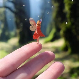 Puede incluir: Una figura de hada en miniatura con vestido naranja y alas de mariposa está de pie sobre un dedo. El hada tiene pelo largo rojo, sobre un fondo borroso de bosque con motas blancas flotantes.