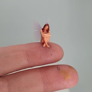 May include: A miniature figurine of a seated figure with reddish-brown hair and translucent, pale wings. The figure is depicted in a curled, seated position with arms wrapped around the legs, showcasing fine details in a very small scale.