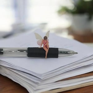 May include: A small fairy figure with delicate wings and a pink dress is perched on a black pen. The pen is placed on a pile of white paper, with a blurred backdrop of a window and a houseplant.