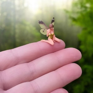 Może przedstawiać: Miniaturowa figurka wróżki ze skrzydłami motyla, w złotym stroju, siedzi na czubku palca. Wróżka ma brązowe włosy i klęczy. Skrzydła są jasnozielone z ciemnymi plamkami. Tło to rozmyty zielony las.