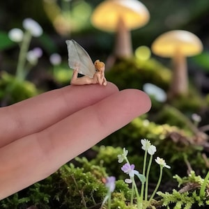 May include: A miniature fairy figurine with blonde hair and translucent wings rests on a fingertip. The fairy is lying down in a forest setting with mushrooms and moss. The scene evokes a whimsical, fantasy theme.