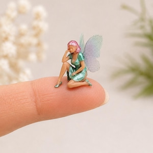 May include: A miniature figurine of a fairy with pink hair, wearing a metallic green dress and matching shoes, featuring iridescent wings. The small figure is posed in a seated position with one hand resting near the face, showing fine details.