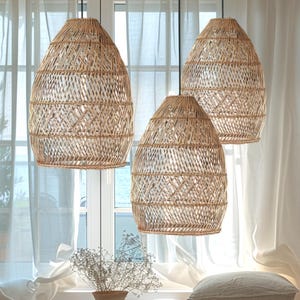 May include: Three woven wicker pendant lights hang from the ceiling in a living room with white curtains and a white sofa. The lights are a natural beige colour and have a geometric pattern.