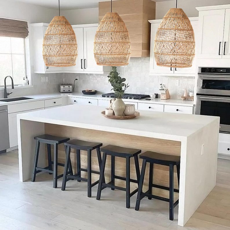 May include: A white kitchen island with a white worktop and four black wooden stools. Three wicker pendant lights hang above the island.