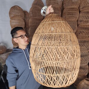 May include: A large, round, woven wicker pendant light fitting. The light is made of natural, light brown rattan and has a diamond pattern. The light is being held by a person.
