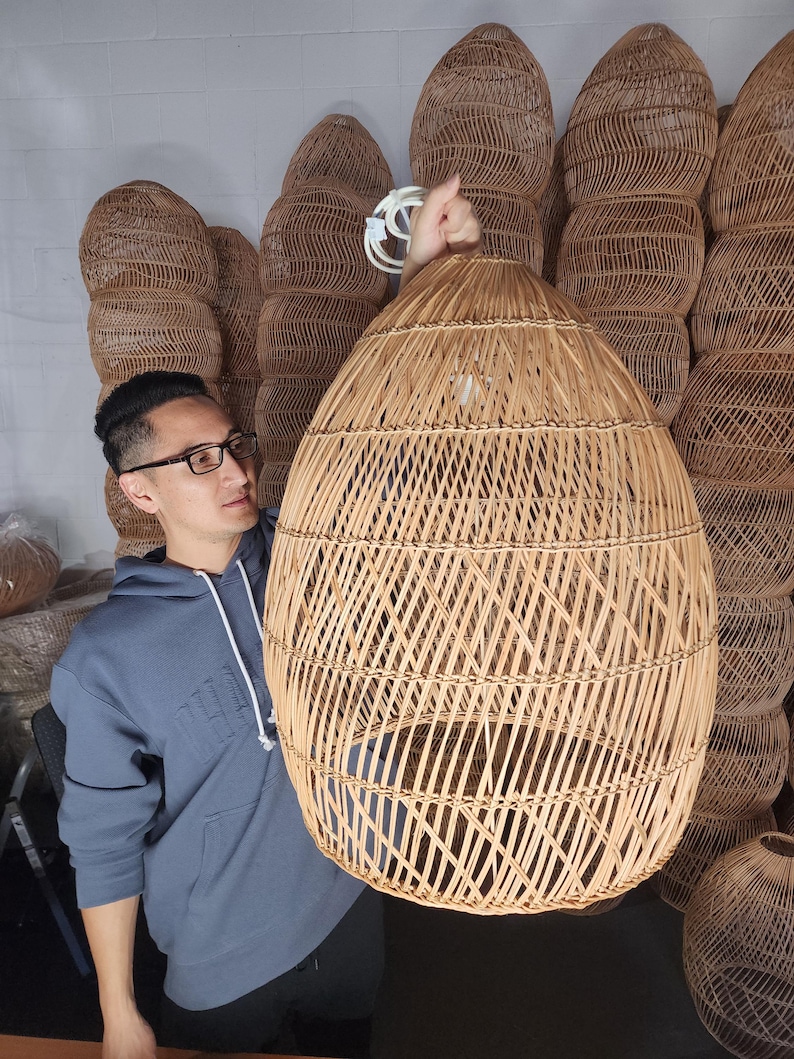 May include: A large, round, woven wicker pendant light fitting. The light is made of natural, light brown rattan and has a diamond pattern. The light is being held by a person.