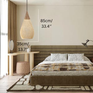 K&ouml;nnte beinhalten: Gewebte Pendelleuchte in Tropfenform, 85 cm hoch und 35 cm im Durchmesser. Die Leuchte hat eine nat&uuml;rliche beige Farbe mit einem braunen Holzaufsatz und h&auml;ngt an einem schwarzen Kabel. Sie befindet sich &uuml;ber einem Bett.