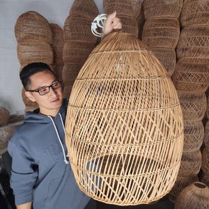 May include: A large, woven wicker pendant light fitting with a natural brown colour. The light is hanging from the ceiling and is being held by a person.