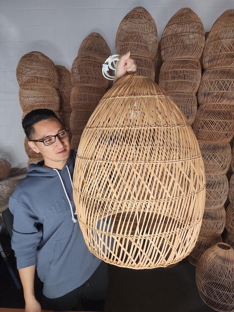 May include: A large, woven wicker pendant light fitting with a natural brown colour. The light is hanging from the ceiling and is being held by a person.