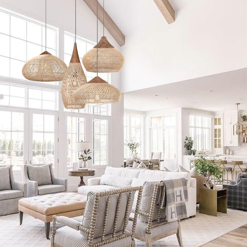 May include: Four woven pendant lights in natural tones hang from the ceiling in a bright living room. The lampshades have various shapes, including a cone and a rounded design. The room features white walls, large windows, and neutral-coloured furniture.