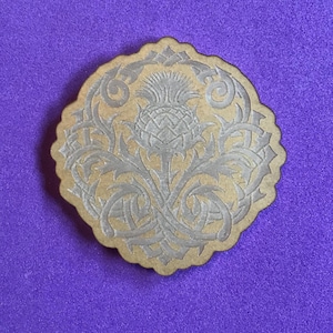 May include: A wooden coaster with a detailed engraving of a thistle flower in a Celtic knot design.
