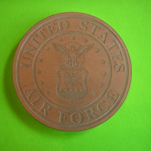May include: A brown wooden coaster with the United States Air Force seal. The seal features a bald eagle with wings spread, a shield with stars, and the words "United States Air Force".