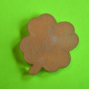 May include: A brown wooden four-leaf clover cutout with the word "lucky" written inside. The cutout is on a green background.