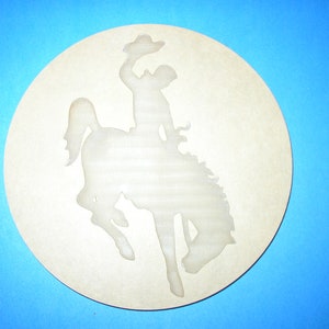 May include: A round, light-colored decorative piece featuring a silhouette of a cowboy riding a bucking bronco. The cowboy is wearing a hat and is in mid-air. The background is a solid blue.