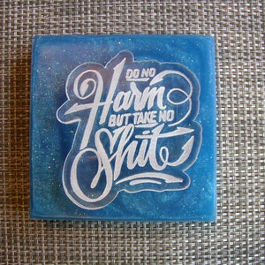 May include: A blue square coaster with a white glitter finish and the text "Do No Harm But Take No Shit" in a stylized font.