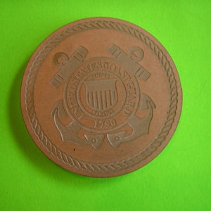 May include: A brown leather coaster with the United States Coast Guard emblem. The emblem features an anchor, a shield with vertical stripes, and the text "United States Coast Guard 1790".