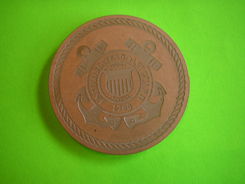 May include: A brown leather coaster with the United States Coast Guard emblem. The emblem features an anchor, a shield with vertical stripes, and the text "United States Coast Guard 1790".