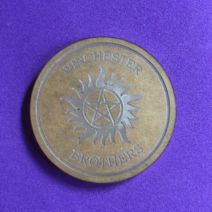 May include: A round, brown wooden coaster with the words "Winchester Brothers" and a stylized symbol of a five-pointed star inside a circle with rays emanating from it.