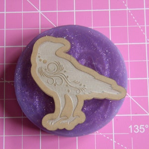 May include: A white, detailed, carved raven design on a purple glitter background. The raven is facing left and has a detailed design on its body.