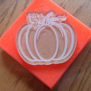 May include: A red rubber stamp with a white outline of a pumpkin with a leaf on top. The stamp is on a wooden surface.