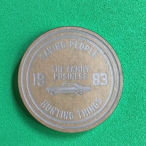 May include: A round, brown wooden coaster with a silver design. The design features the text "Saving People, Hunting Things, The Family Business, 1983".