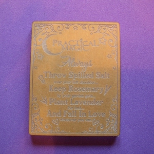 May include: A wooden sign with a decorative border and the text "PRACTICAL MAGIC" in a stylized font. The sign lists several actions to perform for good luck, including throwing spilled salt over your left shoulder, keeping rosemary by your garden gate, planting lavender for luck, and falling in love whenever you can.