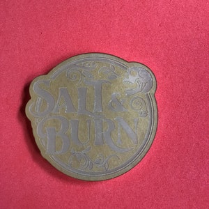 May include: A round, gold-colored coaster with the words "SALT & BURN" in a stylized font. The design includes decorative flourishes and a skull graphic. The coaster is set against a red background.