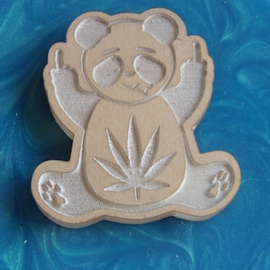 May include: A wooden panda bear figurine with a marijuana leaf on its chest, giving the middle finger with both hands. The figurine is painted white and is sitting on a blue and green surface.