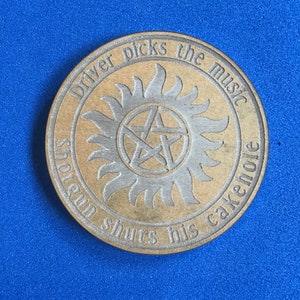 May include: A gold-colored round coin with the text "Driver picks the music shotgun shuts his cakehole" around the edge. In the center is a silver-colored symbol with a five-pointed star and a circle around it.