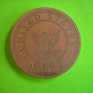 May include: A brown wooden coaster with a raised design of the United States Navy seal. The seal features an eagle with a shield and the words "United States Navy".