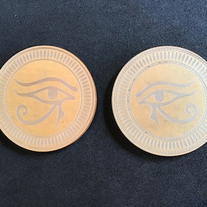 May include: Two round, wooden coasters with a raised, decorative border. Each coaster features a gold-colored, engraved image of the Eye of Horus, an ancient Egyptian symbol.