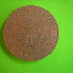 May include: A brown wooden coaster with the United States Army seal. The seal features an eagle with a shield and olive branches. The words "United States Army" are written around the seal.