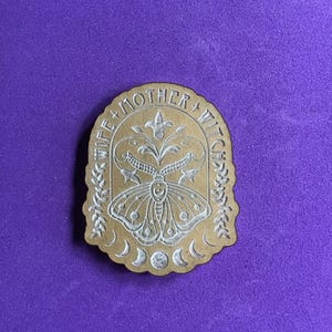 May include: A tan, shield-shaped wooden plaque featuring an engraved moth at the center, surrounded by floral motifs and a lunar cycle at the bottom. The text WIFE + MOTHER + WITCH is engraved in an arch along the top edge of the wooden surface.