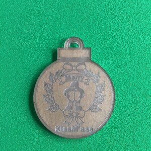 May include: A brown Christmas ornament with a wreath design and the text "Merry Kissmass". The ornament has a loop at the top for hanging. The background is a solid green color.