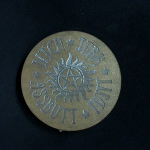 May include: A round, brown wooden coaster with a silver metallic design. The design features a five-pointed star with a circle inside, surrounded by the words "Assbutt Jerk Ditch".