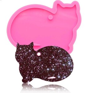 May include: A pink silicone cat-shaped mold with a matching glittery black cat-shaped tag. The mold has a smooth surface, while the tag is covered in sparkling glitter. The tag has a small hole for attaching to a keychain or other item.