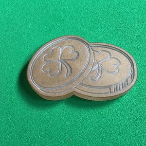 May include: Two wooden coins with a shamrock design, set against a green background. The coins are light brown with a detailed shamrock and border. The image is well-lit, highlighting the texture and design of the coins.