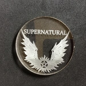 May include: A clear, circular emblem with the word "SUPERNATURAL" etched across the top. Below the text, a detailed design of angel wings and a pentagram is visible. The emblem is set against a dark background.