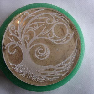 May include: A round, decorative object featuring a detailed tree of life design. The tree is etched in white against a tan background, framed by a silver border and a green outer ring. The design is symmetrical and intricate.