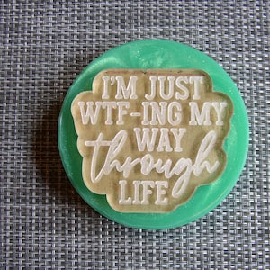 May include: A round, green coaster with a white inscription that reads "I'm just wtf-ing my way through life".