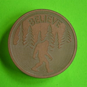 May include: A round, brown wooden coaster with a silhouette of Bigfoot walking through a forest. The text "BELIEVE" is carved above Bigfoot.