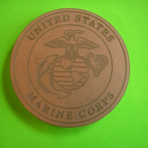 May include: A brown wooden round coaster with the United States Marine Corps emblem. The emblem features an eagle with outstretched wings, a globe, and an anchor.