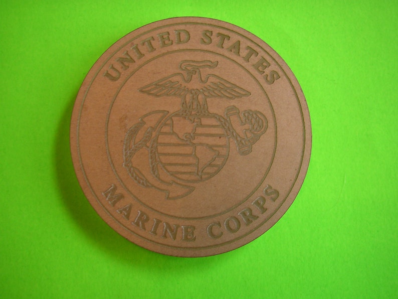 May include: A brown wooden round coaster with the United States Marine Corps emblem. The emblem features an eagle with outstretched wings, a globe, and an anchor.