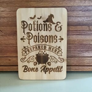 May include: A rectangular wooden sign with the words "Potions & Poisons Prepared Here Bone Appetit" etched into the wood. The sign features a witch's hat, bats, a bubbling cauldron, and skulls, perfect for Halloween or a witch-themed kitchen.