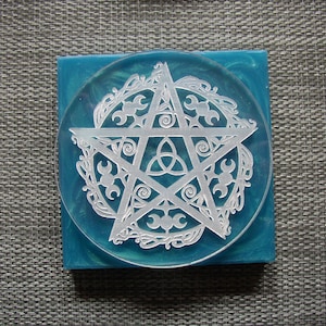 May include: A white pentagram with a triple moon symbol inside, surrounded by a decorative border, is etched into a clear blue resin coaster.