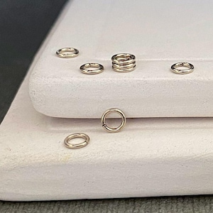 May include: A close-up of a group of small, gold jump rings on a white surface. The rings are arranged in a loose pile, with some of them stacked on top of each other.