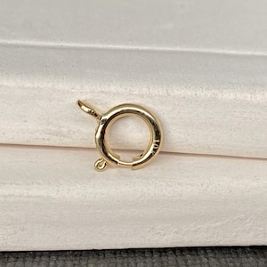以下が含まれることがあります： 金色のスプリングリングクラスプ、ジュエリーコンポーネントが表示されています。 クラスプは円形で、小さなレバーとジュエリーに取り付けるためのループが付いています。 クラスプは金属製で、光沢のある仕上がりです。 背景はニュートラルカラーです。