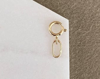 May include: A gold-toned spring ring clasp featuring a small, attached oval jump ring. The circular clasp mechanism includes a tiny lever for opening and closing, and the entire piece is constructed from a polished, metallic gold-colored material.