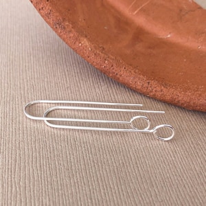 May include: A pair of silver-colored, minimalist earrings. Each earring features a long, curved wire with a small, circular loop at the end. The earrings are displayed on a neutral, textured surface with a terracotta-colored plate in the background.